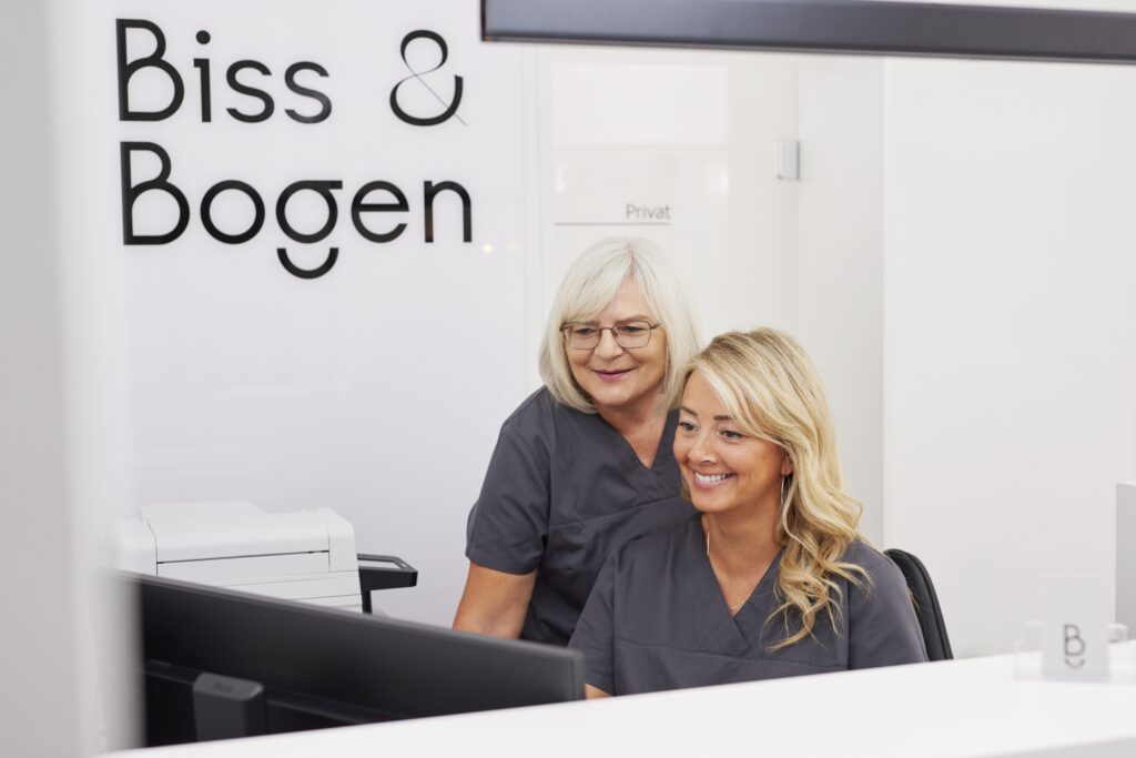 Zwei Mitarbeiterinnen der kieferorthopädischen Fachzahnarztpraxis Biss & Bogen in Ratingen-Lintorf sitzen freundlich lächelnd an einem Empfangscomputer.
