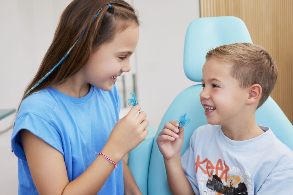 Zwei fröhliche Kinder mit Zahnspangen sitzen in einem Behandlungszimmer der kieferorthopädischen Fachzahnarztpraxis Biss & Bogen in Ratingen-Lintorf und halten blaue Zahnspangen in den Händen.