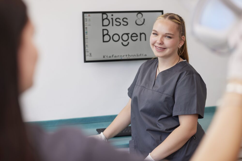 Freundliche Mitarbeiterin in einer kieferorthopädischen Praxis, gekleidet in grauer Arbeitskleidung, lächelt in die Kamera. Im Hintergrund ist ein Bildschirm mit der Aufschrift 'Biss & Bogen Kieferorthopädie' zu sehen.