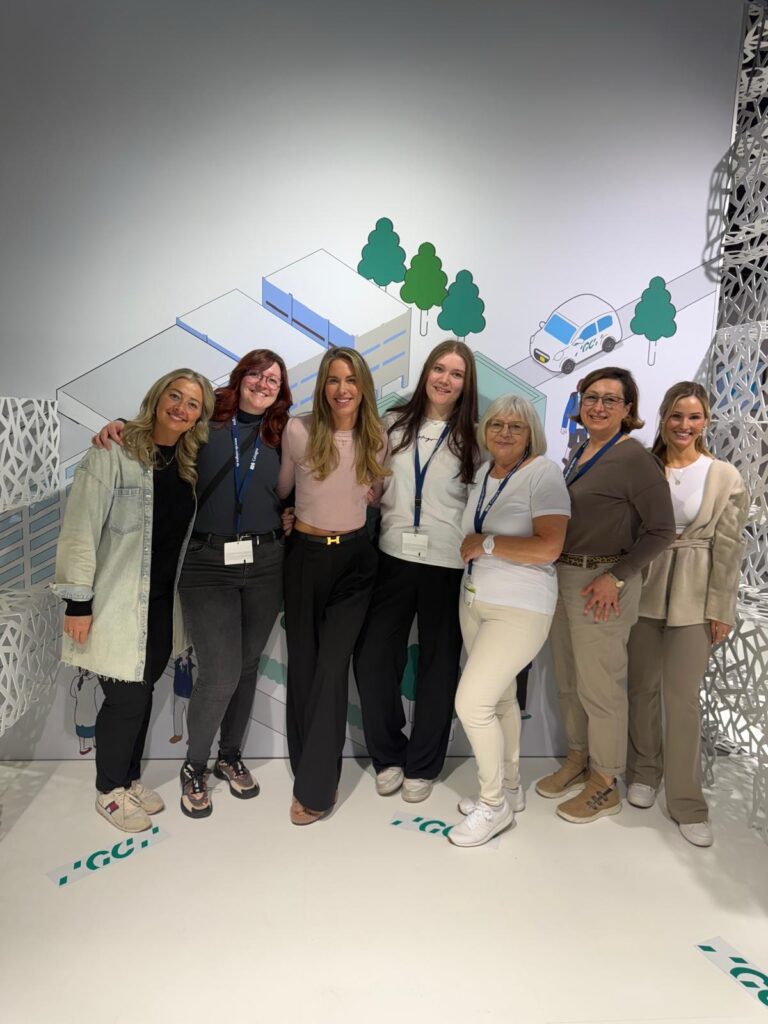 Gruppenfoto des Teams Biss & Bogen auf der IDS 2025 in Köln. Sieben Frauen stehen lächelnd vor einem Messehintergrund mit stilisierter Gebäude- und Naturgrafik.