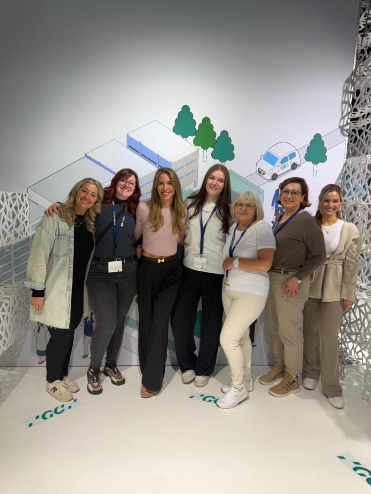 Gruppenfoto des Teams Biss & Bogen auf der IDS 2025 in Köln. Sieben Frauen stehen lächelnd vor einem Messehintergrund mit stilisierter Gebäude- und Naturgrafik.