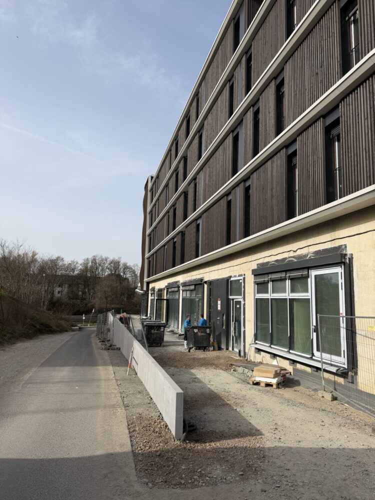 Blick von einem asphaltierten Weg auf ein mehrstöckiges Gebäude mit dunkler Holzlattenfassade in den oberen Stockwerken und hellem Verputz im Erdgeschoss, das noch von einem Baustellenzaun umgeben ist. Im Vordergrund liegen Baumaterialien auf Paletten sowie Mülltonnen, davor stehen zwei Arbeiter in blauer Kleidung vor den großen Fensteröffnungen des Erdgeschosses.