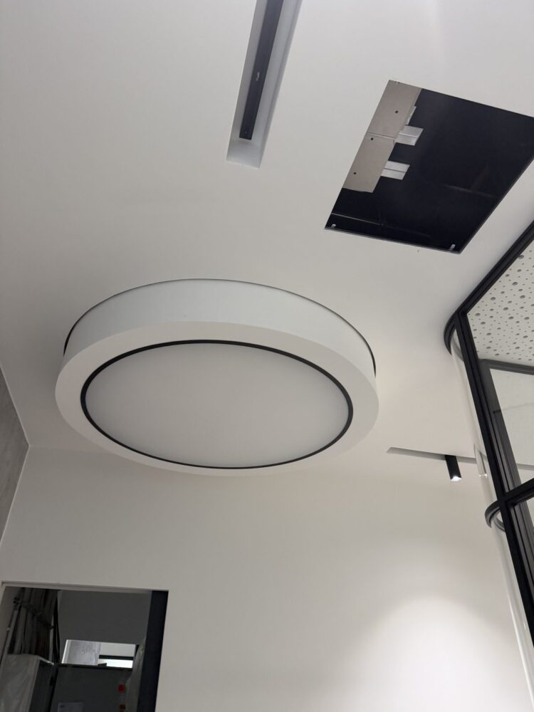 Blick von unten an eine weiße Decke mit einer runden LED-Deckenleuchte mit schwarzem Ring, rechts daneben eine geöffnete Deckenplatte mit sichtbaren Metallprofilen, im Hintergrund ein schwarzer Fensterrahmen mit Glas und eine Türöffnung zu einem angrenzenden Raum.