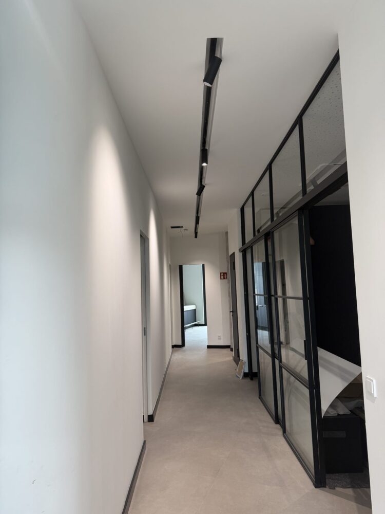 Ein langgestreckter Flur mit weißen Wänden und hellem Fliesenboden, an der Decke eine schwarze Lichtschiene mit mehreren Spotleuchten. Rechts trennt eine schwarze Metall-Glas-Wand einen dahinterliegenden Raum ab, im Hintergrund ist eine offene Tür zu einem weiteren Zimmer mit einem dunklen Einbauschrank zu erkennen.