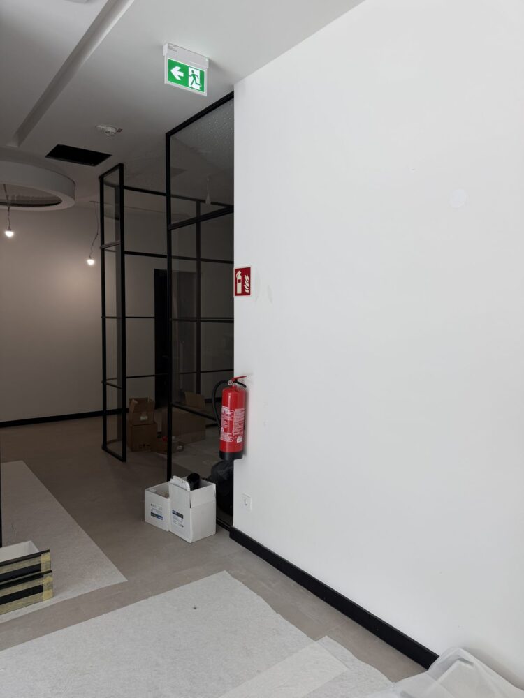 Ein im Bau befindlicher Flurbereich mit weiß verputzten Wänden, an der rechten Wand hängen ein Feuerlöscher und ein Brandschutzschild, darüber ein grün leuchtendes Notausgangsschild. Links führt der Blick durch eine schwarze Glasrahmen-Trennwand in einen weiteren Raum, in dem Kartons auf dem Boden stehen.