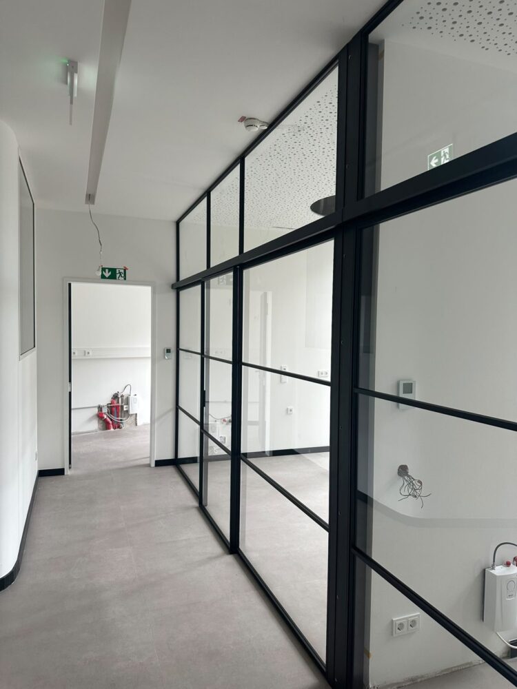 Ein Flur mit grauem Fliesenboden und weissen Waenden, rechts eine raumhohe Glastrennwand mit schwarzen Rahmenprofilen im Rasterdesign, die den Blick in einen dahinterliegenden leeren Raum mit Steckdosen und einem Thermostat an der Wand freigibt. Links im Hintergrund ein Tuerdurchgang mit gruenem Rettungswegschild, davor an der Wand montierte rote Installationsgeraete.
