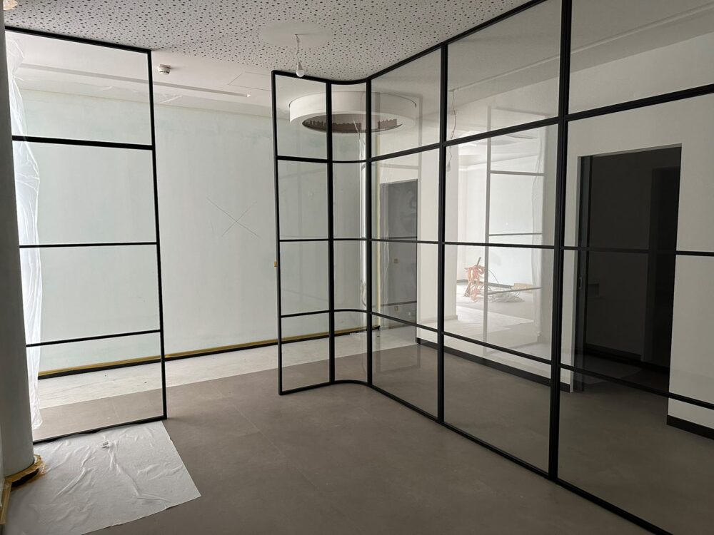 Im Rohbau der Praxis stehen raumhohe Glastrennwände mit schwarzen Metallrahmen, die einen Raum mit gemustertem Akustikdeckel und braunem Fliesenboden unterteilen. An der linken Wand hängen Schutzfolien, der Boden ist teilweise mit Malervlies abgedeckt, im Hintergrund sind weitere Räume mit Türöffnungen und Baumaterial zu erkennen.