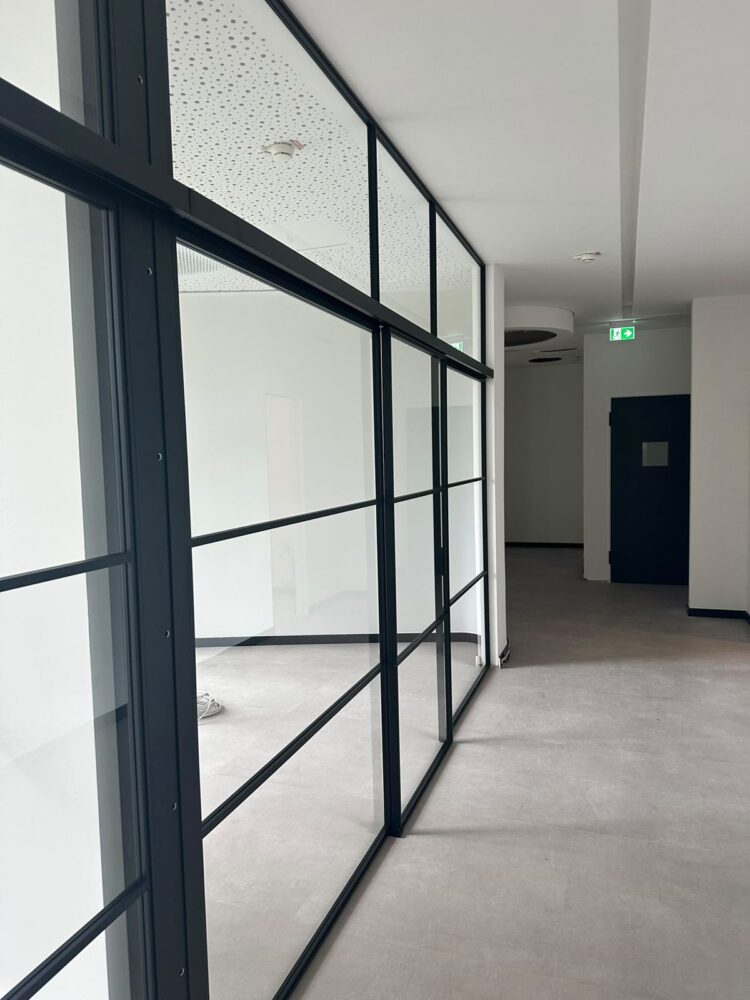 Foto eines noch im Bau befindlichen Flurs mit hellgrauem Boden und weissen Waenden. Links verlaeuft eine raumhohe Glaswand mit schwarzen Rahmenprofilen, die einen leeren Raum mit gepunkteter Akustikdeckendecke abgrenzt, im Hintergrund eine dunkle Tuer und ein gruenes Fluchtwegschild.