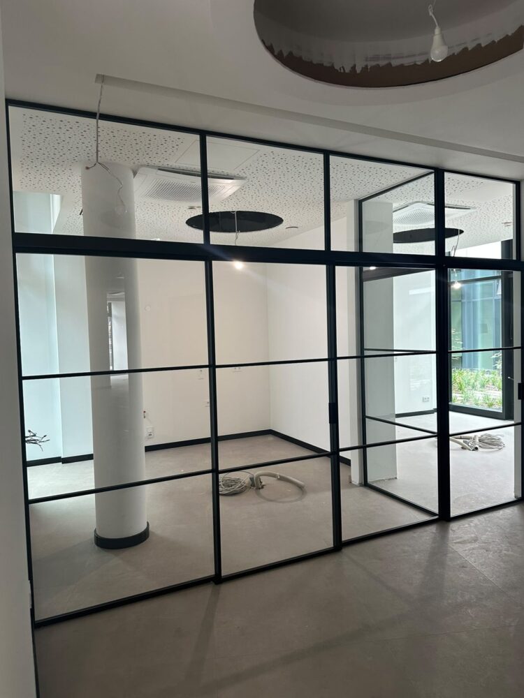 Im Rohbau sichtbare Trennwand aus schwarz gerahmten Glaselementen zwischen zwei leeren Raumbereichen mit grauem Fliesenboden, im Hintergrund eine weiße Säule und lose verlegte Kabel auf dem Boden, die Decke ist teilweise mit Akustikplatten verkleidet und zeigt eine offene Montageöffnung mit herabhängenden Kabeln.