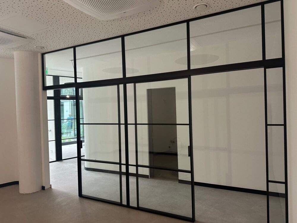Im Rohbau der Praxis steht eine raumhohe Trennwand aus Glas mit schwarzem Metallrahmen, unterteilt in mehrere Segmente mit einer Schiebetuer. Im Hintergrund ist ein Flur mit einer weiteren Tuer sowie links ein Ausgang mit Blick nach draussen zu erkennen, die Decke zeigt Lochplatten und Lueftungselemente.