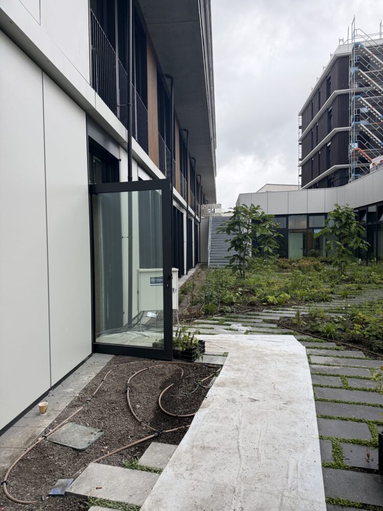 Blick entlang der weißen Fassade eines zweigeschossigen Gebäudes mit offener Glastür im Erdgeschoss, davor liegen Bewässerungsschläuche auf offener Erde. Im Hintergrund führt ein gepflasterter Weg durch eine bepflanzte Grünfläche zu einem weiteren Gebäudetrakt, rechts ist ein Nachbargebäude mit Baugerüst zu sehen.