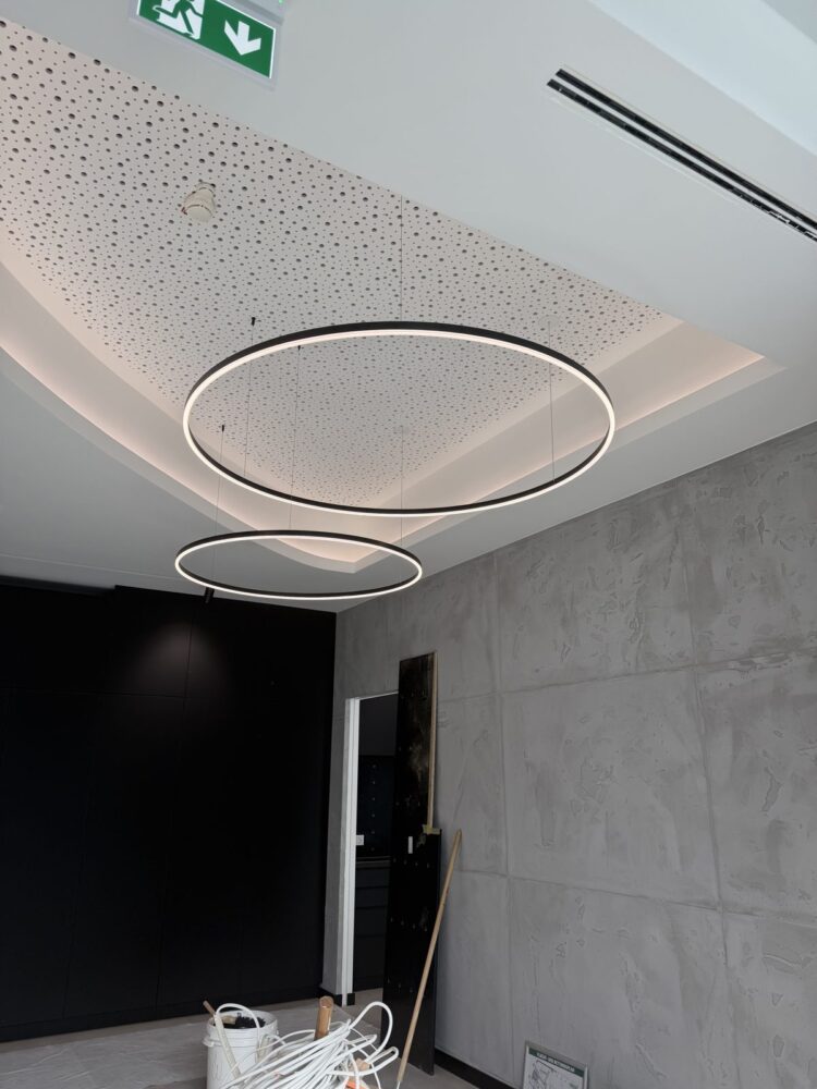 Blick an eine Decke im Rohbau mit zwei ringförmigen schwarzen LED-Hängeleuchten unterschiedlicher Größe, die von einer perforierten Deckenverkleidung herabhängen. Im Hintergrund sind eine grau strukturierte Wand, ein schwarzer Einbauschrank, ein Rettungswegschild sowie Baumaterial und Kabel auf dem Boden zu sehen.
