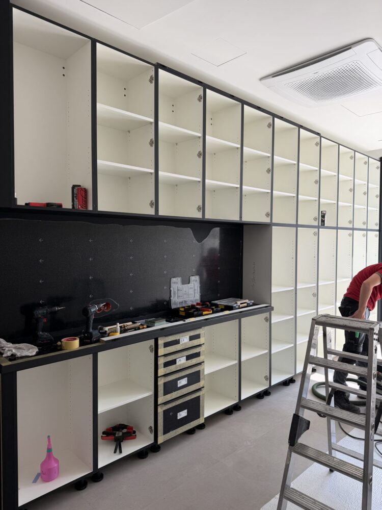 Ein Handwerker steht auf einer Leiter und arbeitet an einer hohen weißen Regalwand mit vielen leeren Fächern, im Vordergrund steht eine schwarze Unterschrankreihe mit offenen Fächern und Schubladen, auf der Arbeitsplatte liegen Akkuschrauber, Werkzeugkoffer und Klebeband.