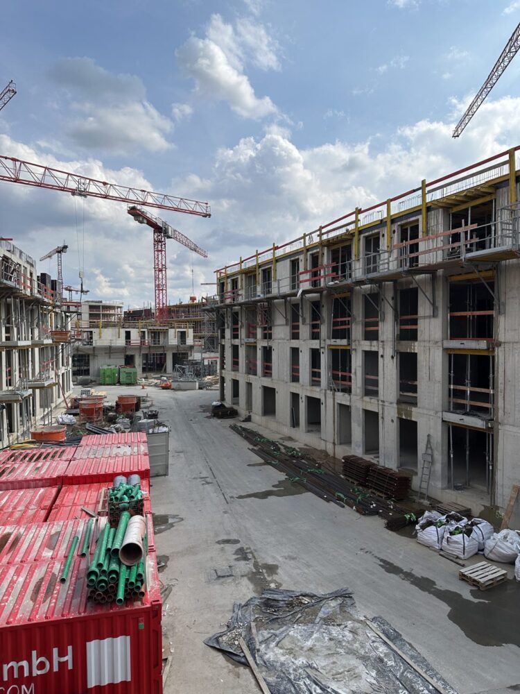 Blick auf mehrere mehrstoeckige Rohbauten aus Beton mit offenen Fensteroeffnungen und Deckenschalungen, im Hintergrund mehrere rote Turmdrehkrane vor bewoelktem Himmel. Im Vordergrund stehen rote Materialcontainer mit gruenen und grauen Rohren sowie am rechten Bildrand gebuendelte Bewehrungsstaebe und weisse Saecke auf dem Baustellenboden.