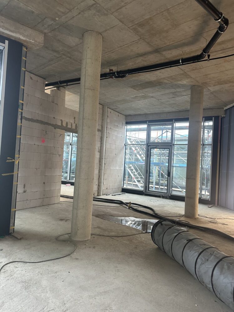 Blick in einen unverputzten Rohbauraum mit zwei runden Betonstützen, gemauerten Wänden aus Kalksandstein und einer Fensterfront mit Terrassentür, davor ist außen ein Baugerüst zu sehen. Auf dem Betonboden liegen ein grauer Lüftungsschlauch, mehrere Kabel und Schläuche sowie eine Pfütze, an der Decke verlaufen Rohrleitungen.