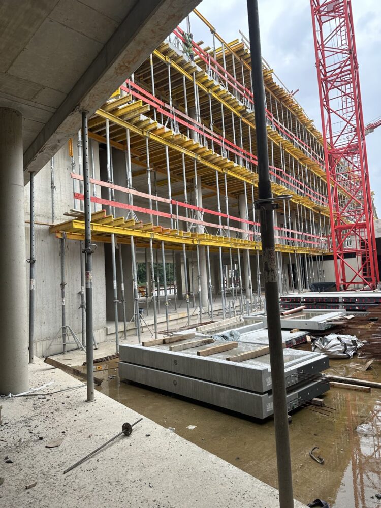 Blick durch einen Betonrohbau auf ein Gerüst mit gelb-roter Deckenschalung über mehrere Stockwerke, rechts ein roter Baukran vor bewölktem Himmel. Im Vordergrund stehen Stützstempel, gestapelte Betonfertigteile und Bewehrungsstahl auf nassem Untergrund.