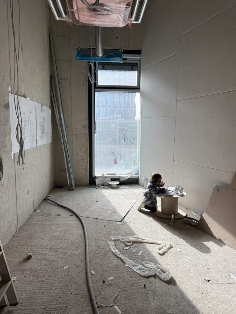 Ein Rohbauraum mit unverputzten Betonwaenden, Kabeln, Bauschutt und Kartonresten auf dem Boden, im Hintergrund ein grosses Fenster mit Blick auf ein Nachbargebaeude mit der Aufschrift Essential. Vor dem Fenster sitzt ein Kind auf dem Boden an einer umgedrehten Kabeltrommel und malt oder schreibt auf einem Blatt Papier.