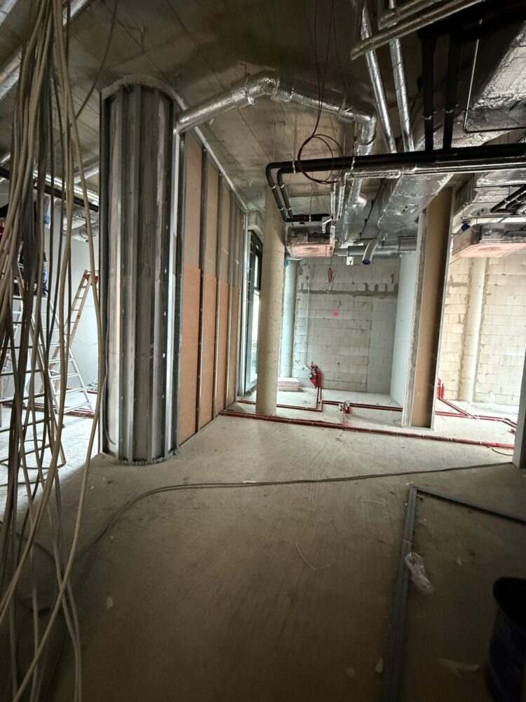 Unfertiger Praxisraum im Rohbau mit sichtbarer Betondecke, Metallprofilen fuer Trockenbauwaende und einer runden Saeulenverkleidung im Vordergrund links. An der Decke verlaufen isolierte Lueftungsrohre und Kabel, im Hintergrund sind gemauerte Waende, eine Betonstuetze und am Boden verlegte rote Rohrleitungen zu sehen, links stehen zwei Leitern.
