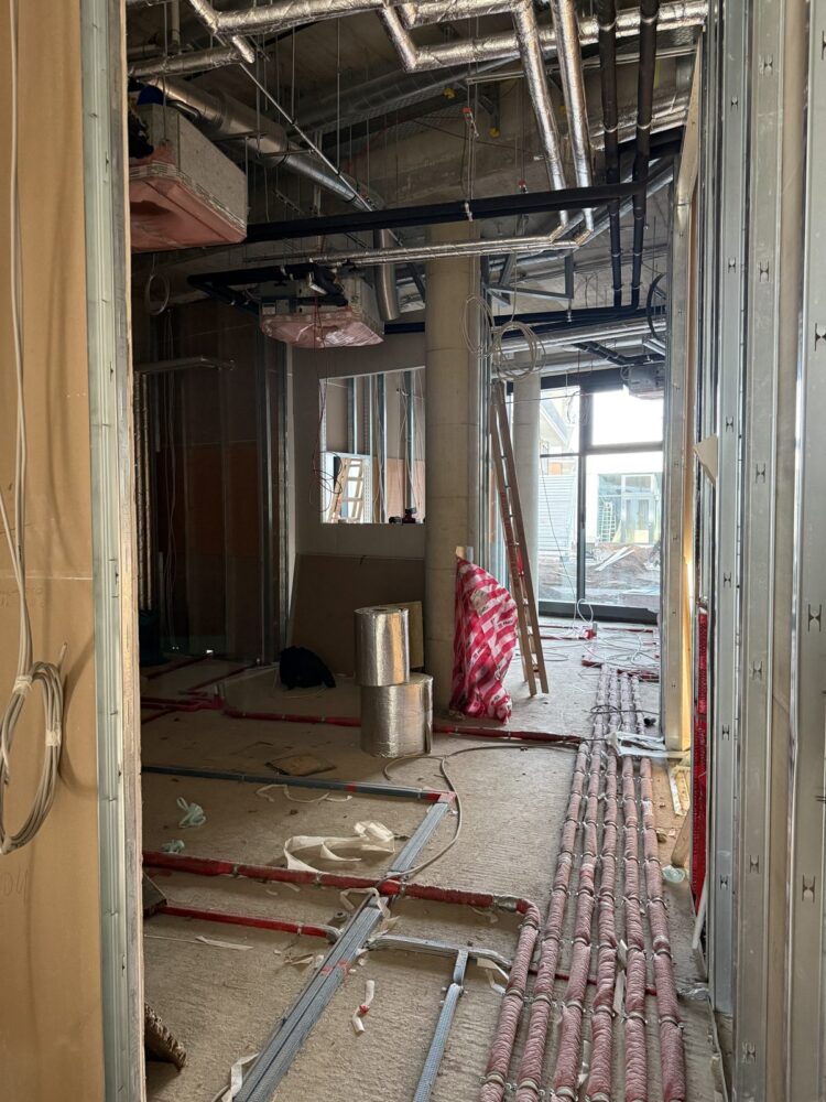 Blick durch einen Rohbaukorridor mit sichtbaren Metallständerwänden auf einen Raum im Ausbauzustand. An der Decke verlaufen zahlreiche Lüftungsrohre und Kabel, im Boden liegen gebündelte rote und silberne Leitungen sowie eine Aluminiumrolle, im Hintergrund stehen eine Leiter und ein Betonpfeiler vor einer Fensterfront.