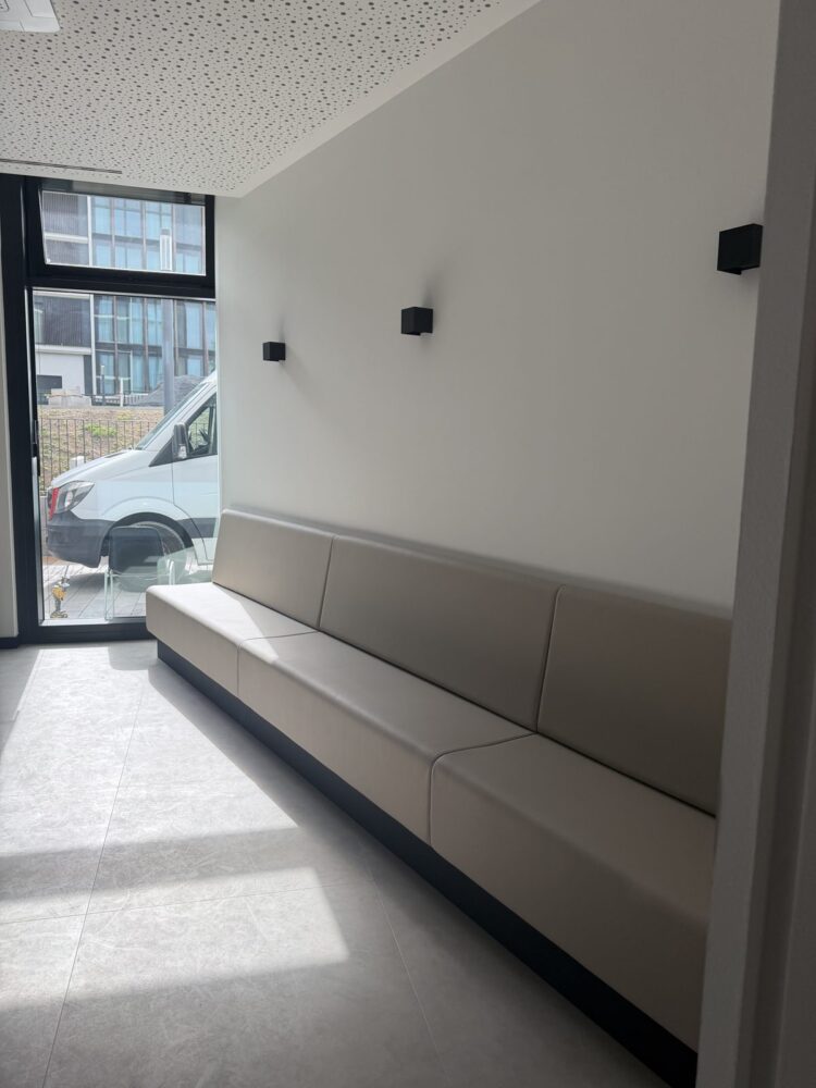 Blick in einen Flur mit gepolsterter Sitzbank entlang der Wand, darüber drei quadratische Wandleuchten aus schwarzem Metall. Links führt eine Glastür mit Blick auf einen weißen Transporter und ein Nachbargebäude nach draußen, der Boden ist mit hellgrauen Fliesen belegt.