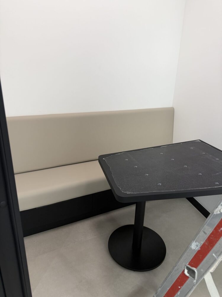 In einer Raumecke steht eine gepolsterte Sitzbank in Beige mit dunkler Sockelblende, davor ein kleiner Tisch mit schwarzer Tischplatte und rundem Metallfuss, dessen Oberflaeche noch mit Schutzfolie und Markierungspfeilen versehen ist. Rechts im Bild lehnt eine Trittleiter mit rot-weissen Streifen an der Wand.