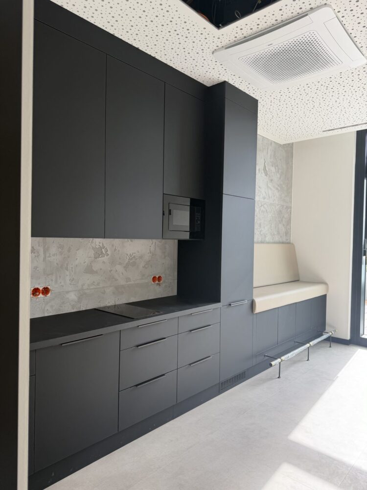Eine im Bau befindliche Küchenzeile mit dunkelgrauen Schrankfronten, integrierter Mikrowelle und Kochfeld auf schwarzer Arbeitsplatte, daneben eine gepolsterte Sitzbank in Creme. An der Decke hängt ein Klimagerät, die Wand hinter der Küche zeigt eine unverputzte Betonoptik mit freiliegenden Steckdosen.