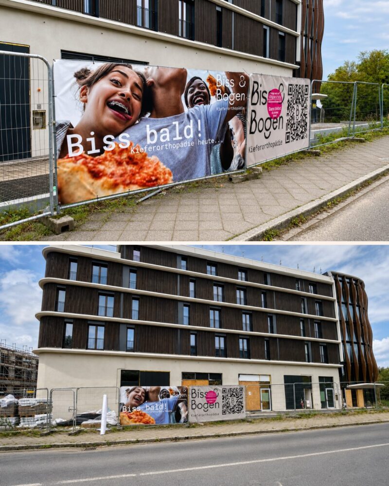 Zwei Fotos zeigen an einem Bauzaun befestigte Werbebanner der Praxis Biss & Bogen vor einem mehrstöckigen Gebäude mit Holzlattenfassade. Auf dem großen Banner sind zwei lachende junge Frauen mit Zahnspangen und ein Pizzastück abgebildet, daneben ein zweites Banner mit Logo, QR-Code und dem Schriftzug Kieferorthopädie; im Hintergrund sind die noch verschlossenen Fensteröffnungen und Bauelemente des Erdgeschosses zu erkennen.